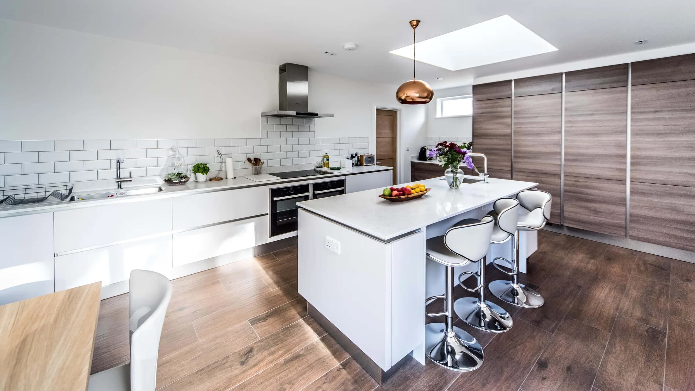 Kitchen with white handleless units, island with bar stools, metro-tile splashback and a large rooflight